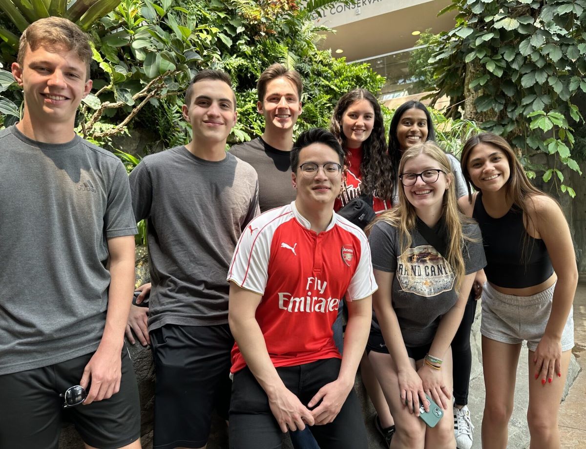 A group of nine young adults pose together in an indoor garden or conservatory filled with lush green plants and vines. Some are standing while others are sitting or leaning on a low stone ledge. Everyone is smiling and dressed casually in t-shirts, athletic wear, and shorts. One person in the center wears a bright red soccer jersey, which stands out against the greenery. The space is well-lit with natural light, and the overall mood feels friendly, relaxed, and social.