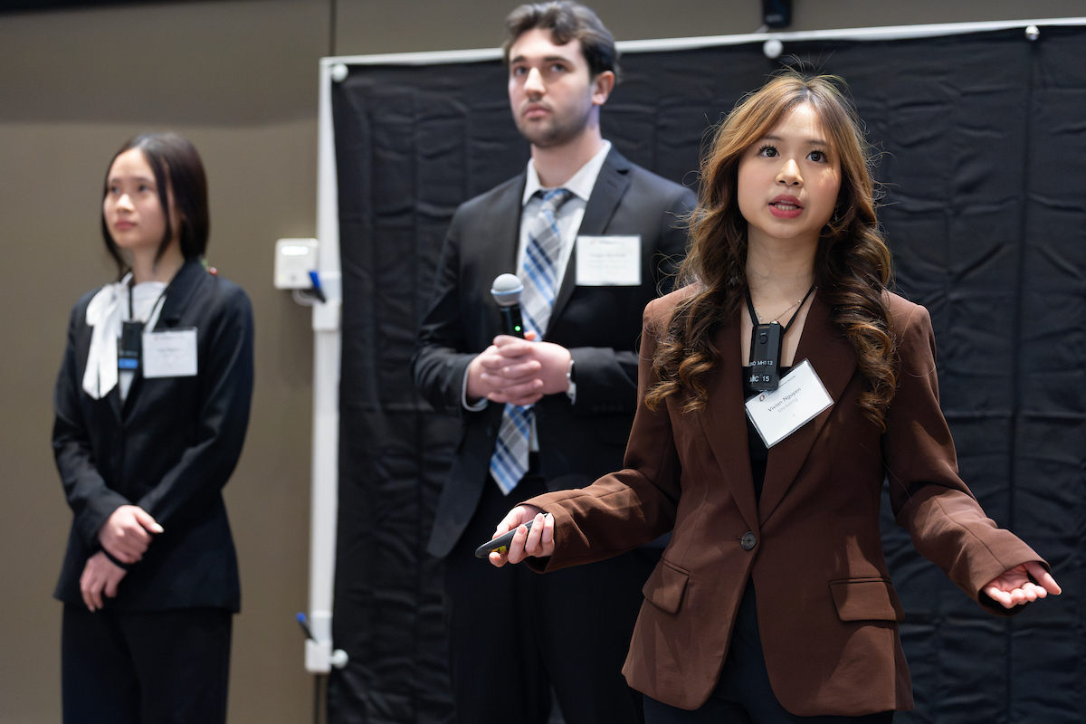 A team of three young professionals stands presenting in a conference room. A woman in a brown blazer speaks in the foreground while holding a small remote. Behind her, a man in a suit holds a microphone, and another woman in a black suit stands to the side. All three wear name badges and appear engaged in a formal presentation.
