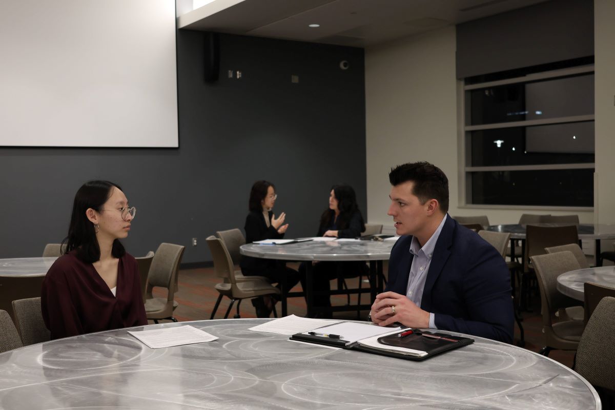 Two people in business attire sit across from each other at a round table in a modern meeting space, engaged in conversation. Papers and a portfolio folder lie on the table between them. In the background, two other pairs of people sit at separate tables talking, suggesting a networking event or interview setting.