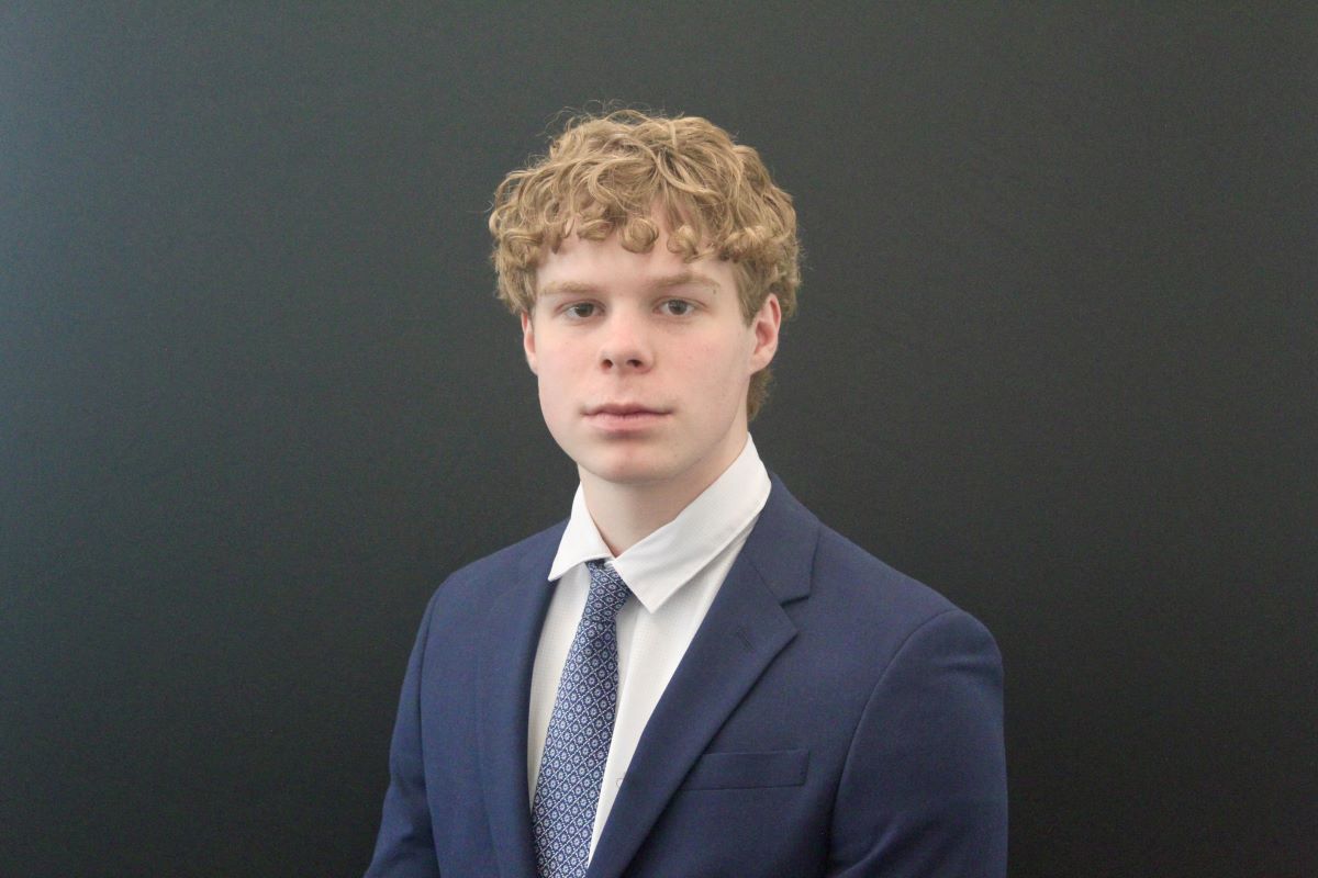 A young man with curly blond hair poses against a dark background, wearing a navy blue suit jacket, white dress shirt, and patterned tie. He faces the camera with a neutral expression.