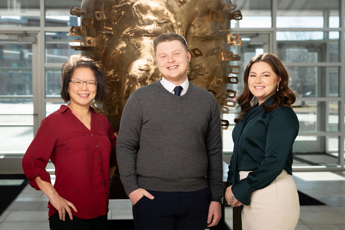 Three colleagues stand side by side in a bright, modern indoor atrium with large windows. They are smiling and dressed in professional business attire. Behind them is a large circular bronze sculpture mounted on a stand.