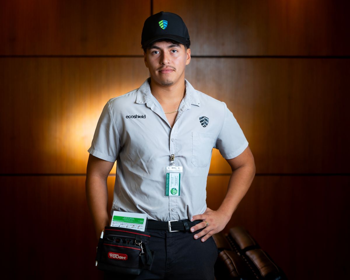 A man wearing an EcoShield uniform and cap stands confidently with one hand on his hip, a tool pouch at his side, in an indoor setting with a warm-toned background.