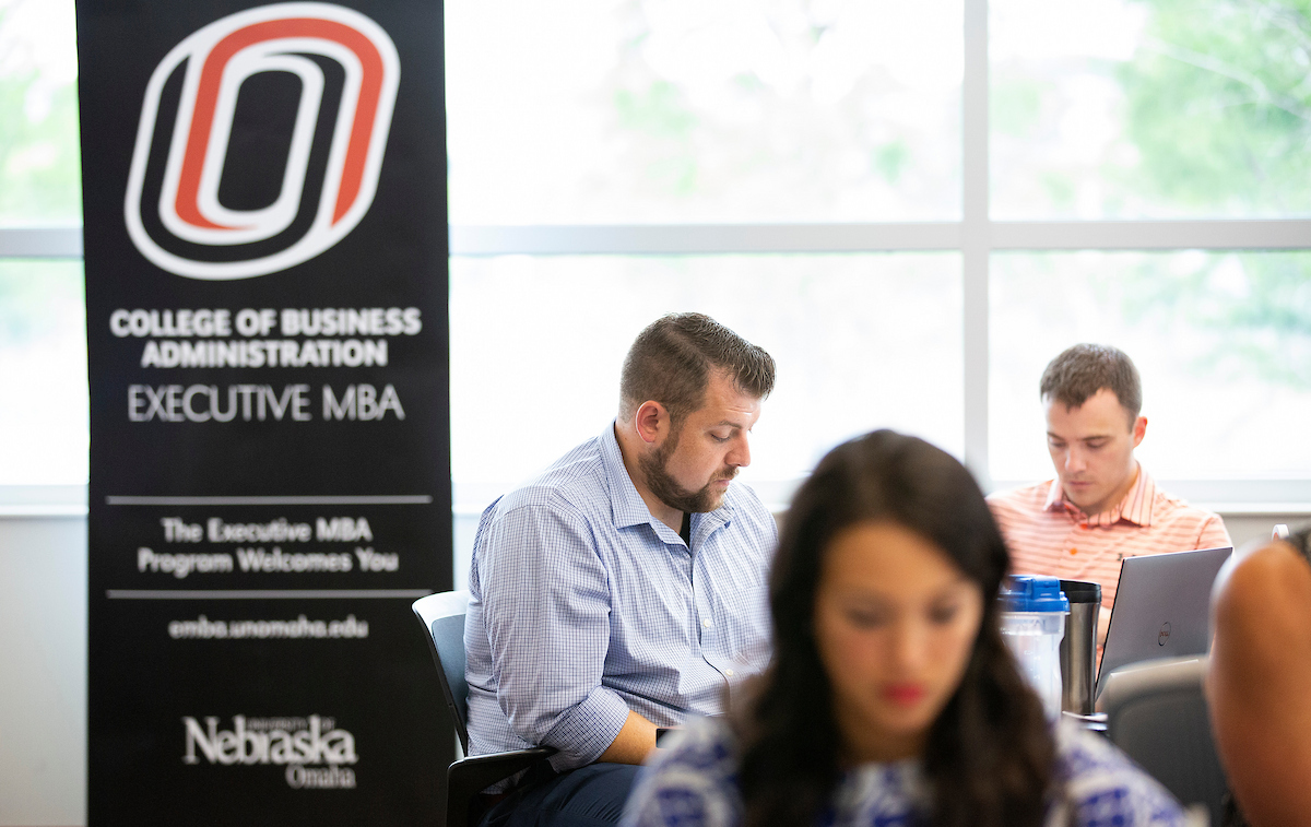 Students sit in a classroom working on laptops and notes, with a University of Nebraska at Omaha College of Business Administration Executive MBA banner visible in the background.