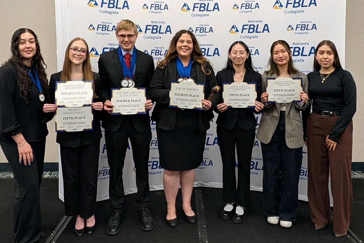 A group of students stand on a stage in front of a Nebraska FBLA Collegiate backdrop, holding certificates and medals for their competition placements, dressed in professional attire and smiling.