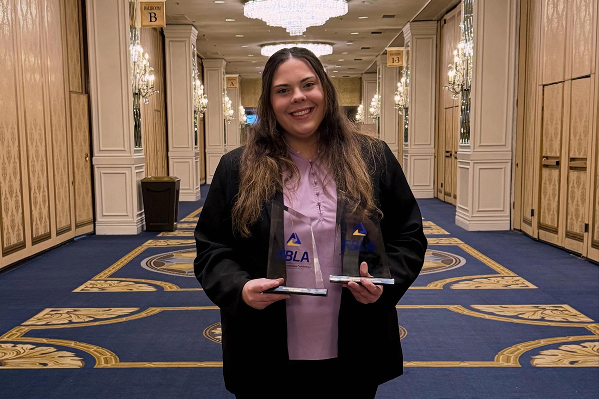 A smiling person stands in the center of an elegant hotel ballroom hallway, holding two glass award trophies. They are wearing a light purple blouse and a dark blazer. Crystal chandeliers hang from the ceiling, and decorative blue-and-gold carpeting extends down the corridor. Signs for ballroom meeting rooms are visible in the background. The person appears to be posing proudly after receiving recognition at a professional or academic event.