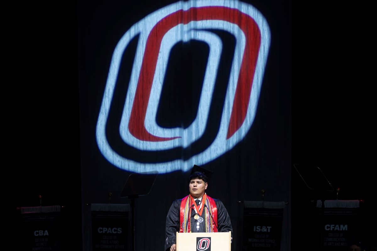 A University of Nebraska at Omaha graduate stands at the commencement podium wearing a cap, gown, honor cords, medals, and a red stole. A large illuminated UNO logo is projected behind the speaker, with college banners visible on either side of the stage.