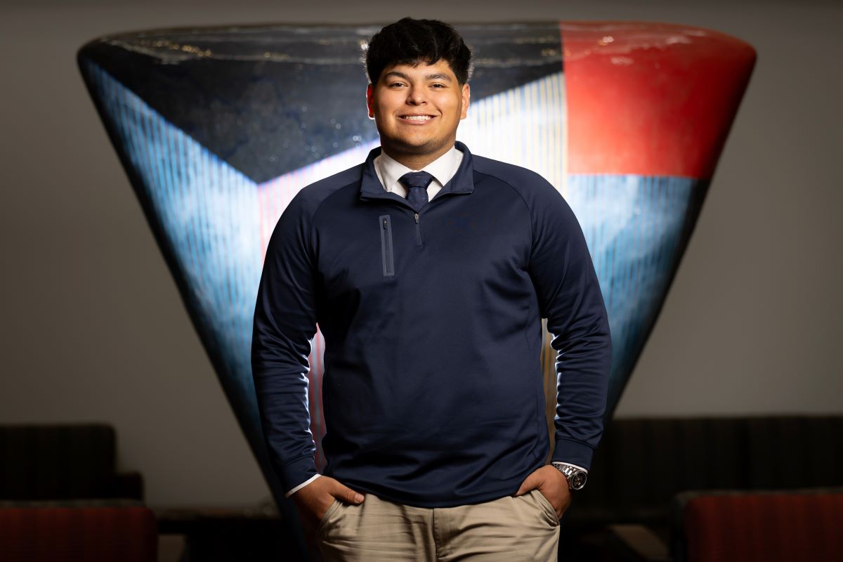 A young man with dark hair, wearing a navy pullover, white shirt, and tie, stands smiling with his hands in his pockets.