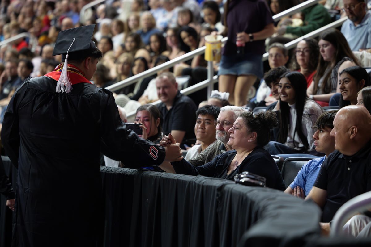 A University of Nebraska at Omaha graduate in a cap and gown reaches into the audience to clasp hands with a family member during commencement, as relatives smile and watch from the front row of a packed arena.