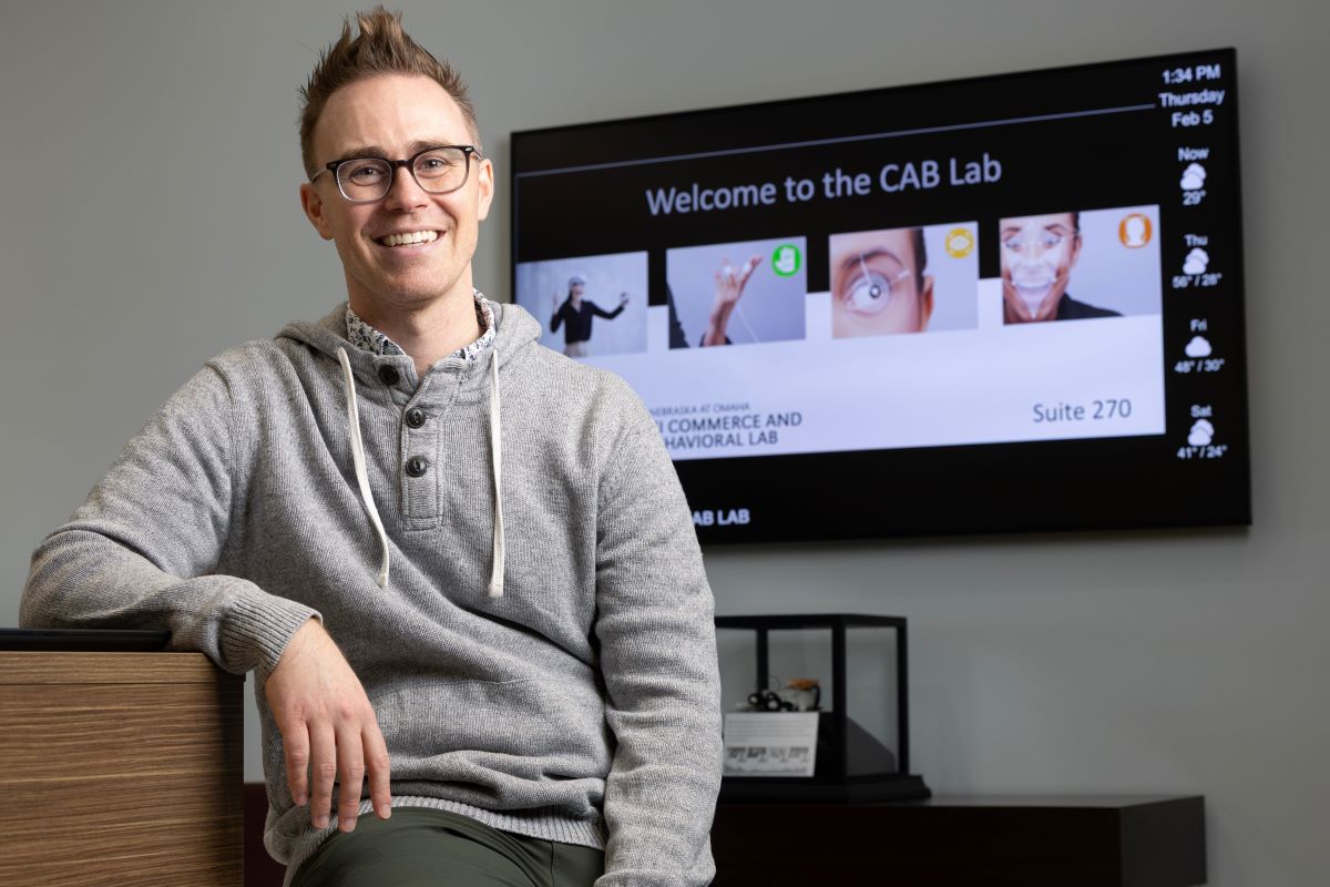 A man wearing glasses and a gray sweater smiles while leaning against a counter in the UNO Commerce and Behavioral Lab (CAB Lab), with a digital display visible behind him.