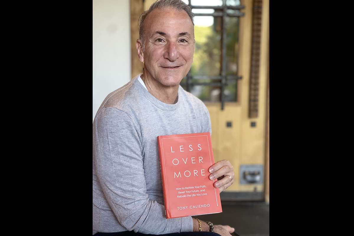 A smiling man in a gray sweater holds a copy of his book, Less Over More, while seated indoors.