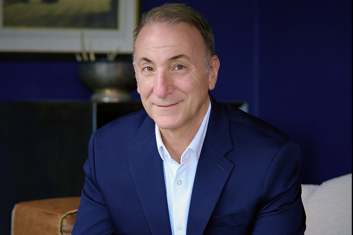A man in a navy suit and white shirt sits on a leather sofa, smiling at the camera, with a dark blue wall in the background.
