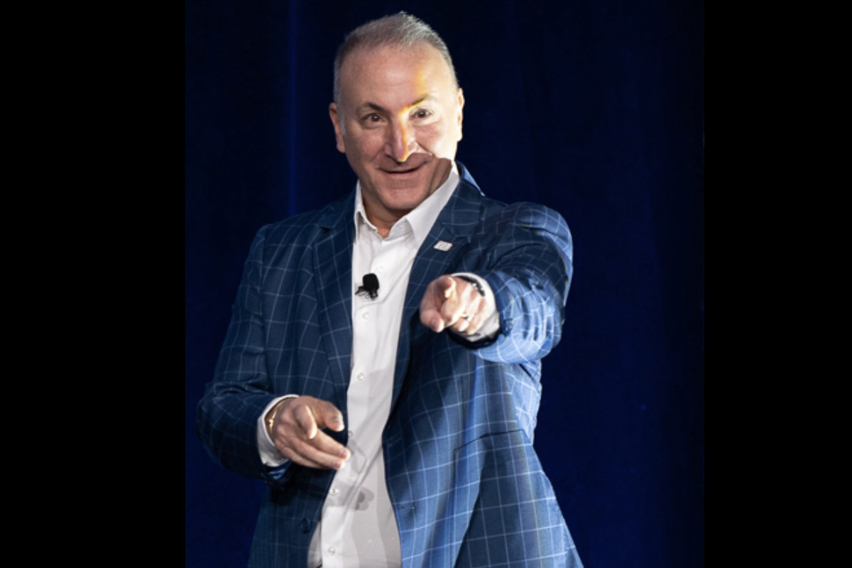 A man in a blue plaid blazer speaks onstage, smiling and pointing toward the audience against a dark blue backdrop.