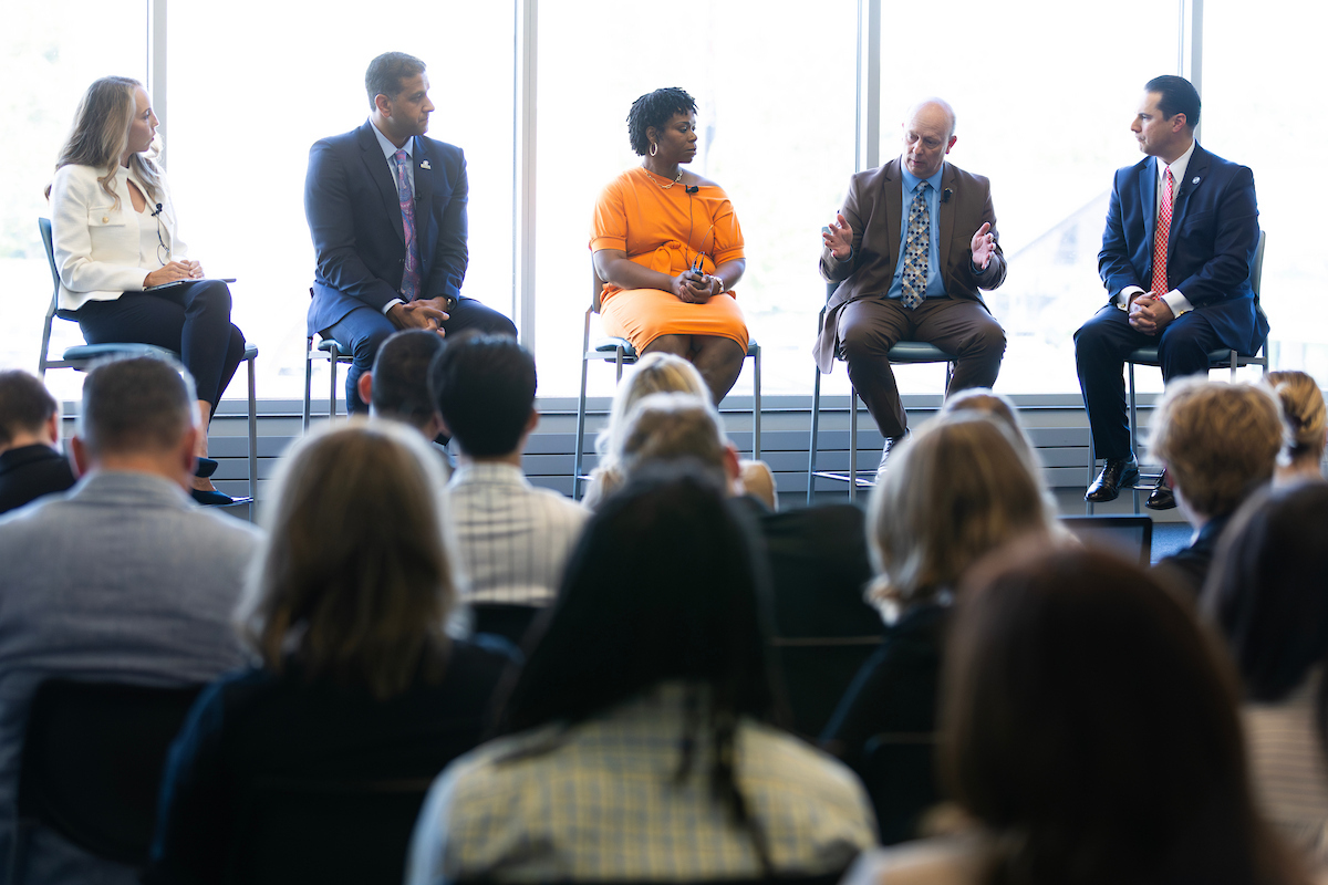 5 individuals sitting in chairs at the front of a room speaking to an audience sitting in chairs.