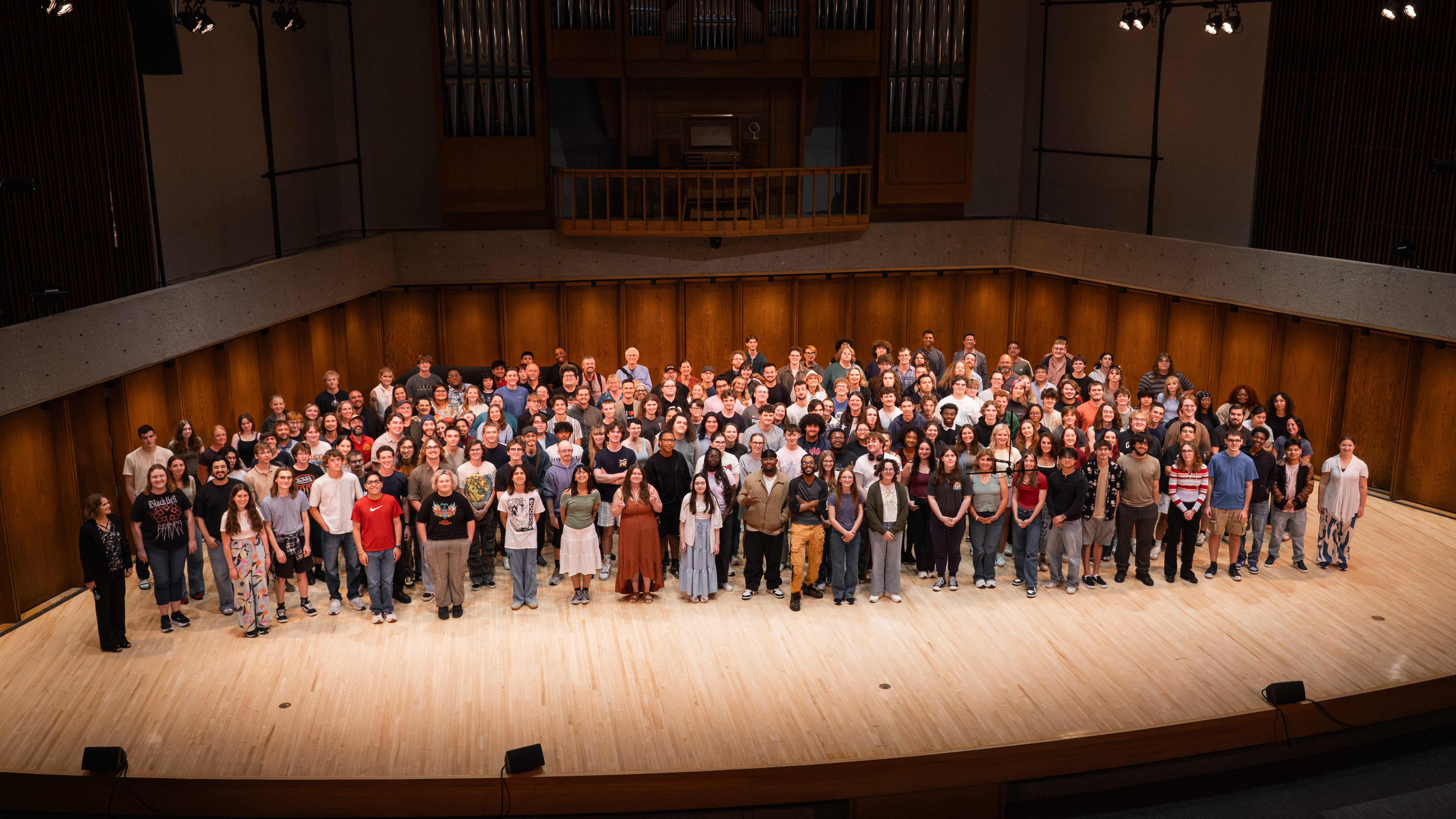 Large group photo of students and staff standing on a wood stage in a concert hall, with an organ loft behind them.