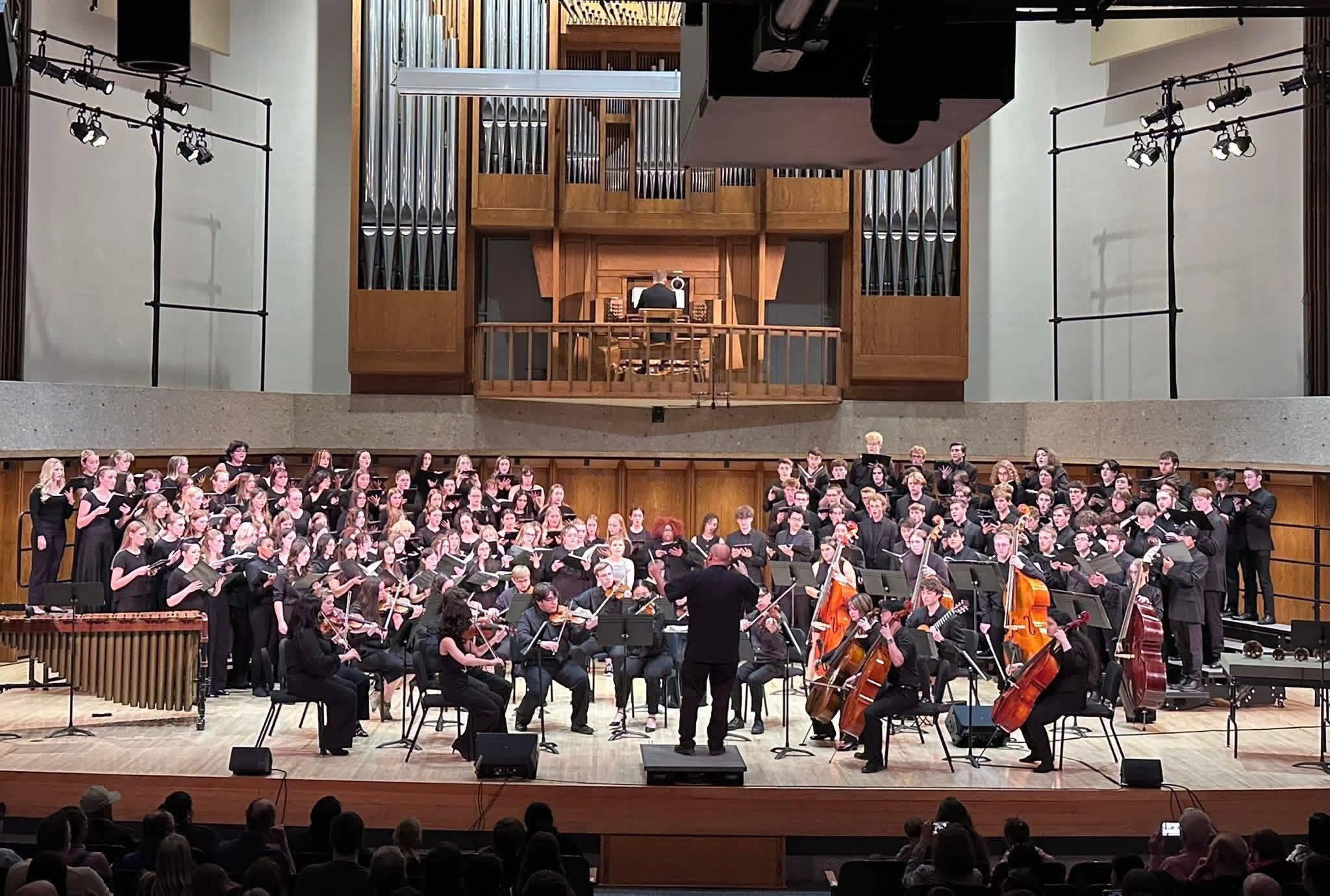 Group of students lined up on a stage, singing and moving in unison during a rehearsal in a modern auditorium.