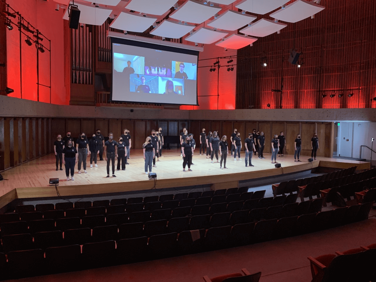 The UNO Concert Choir standing on a wooden stage with red lighting. All students are wearing jeans, black tee shirts, and masks.