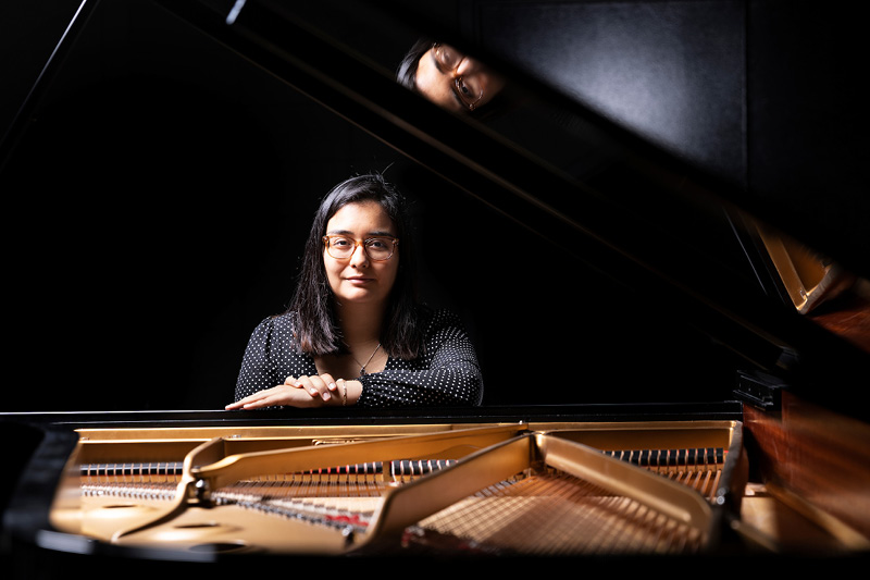 Senior Aida Soria sitting at a piano smiling. Her face is reflected on the open piano lid.