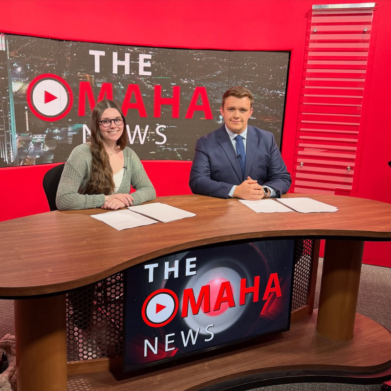 students smiling at a desk with a red background and monitors showing the words omaha news