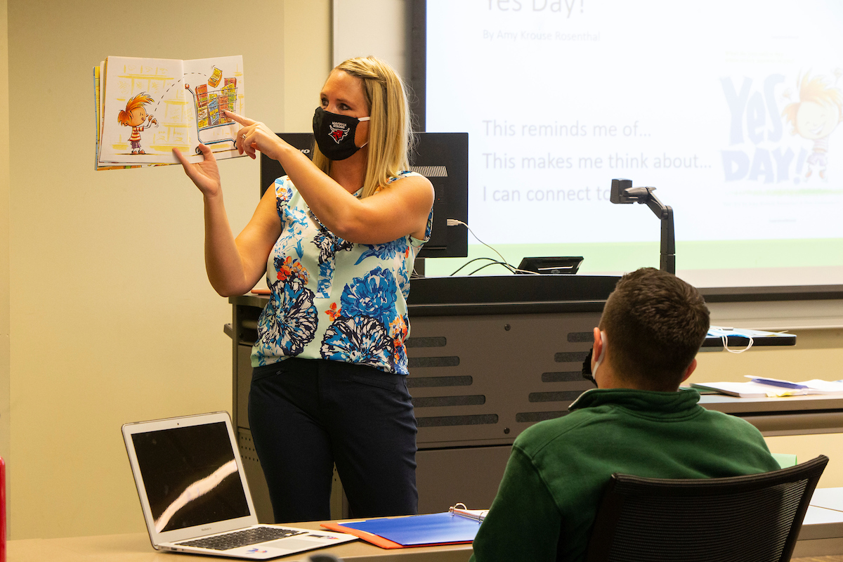 Dr. Jennifer Lemke stands at the front of a classroom holding a children's book