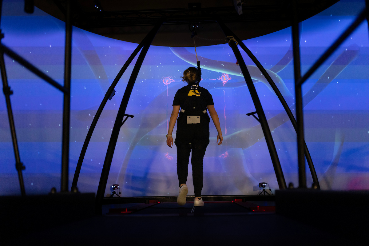 person in a harness walking on a treadmill in front of a virtual reality projector screen