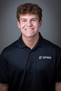 a person with short brown hair smiling wearing a black polo.