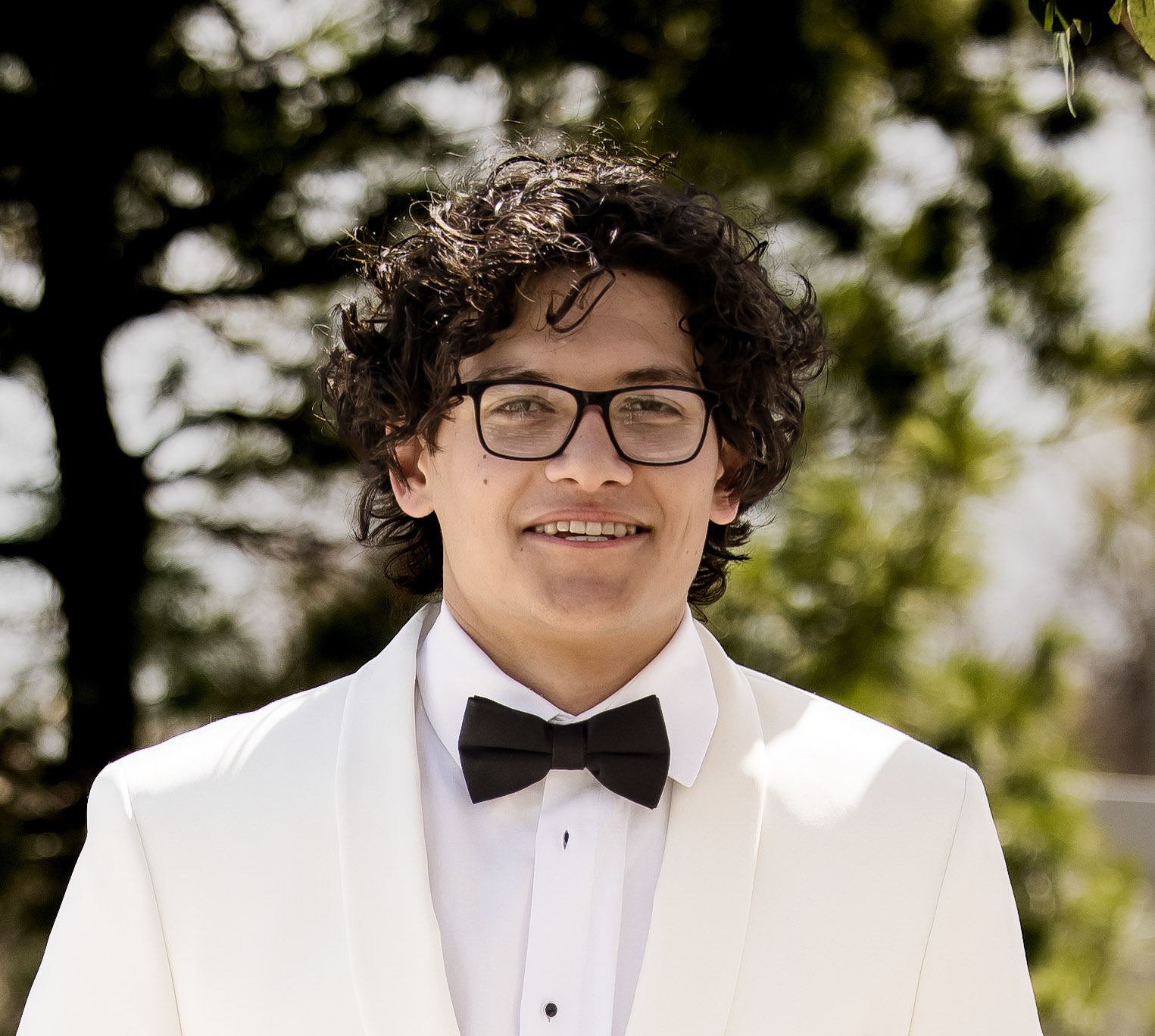 a person smiling with short brown curly hair wearing glasses and a white tuxedo.