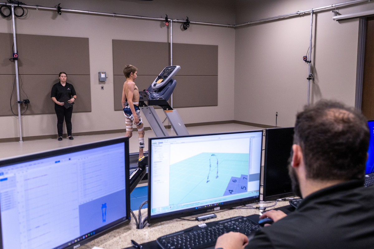 a child walks on force plates with markers while one person observes and another looks at data on a computer