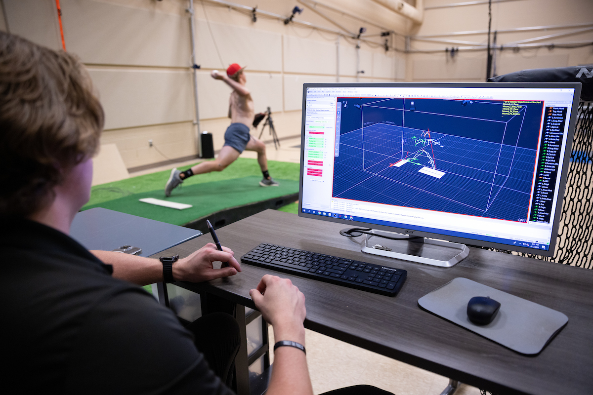 a person sits at a computer observing a person pitching a baseball to assess the biomechanical data on the computer