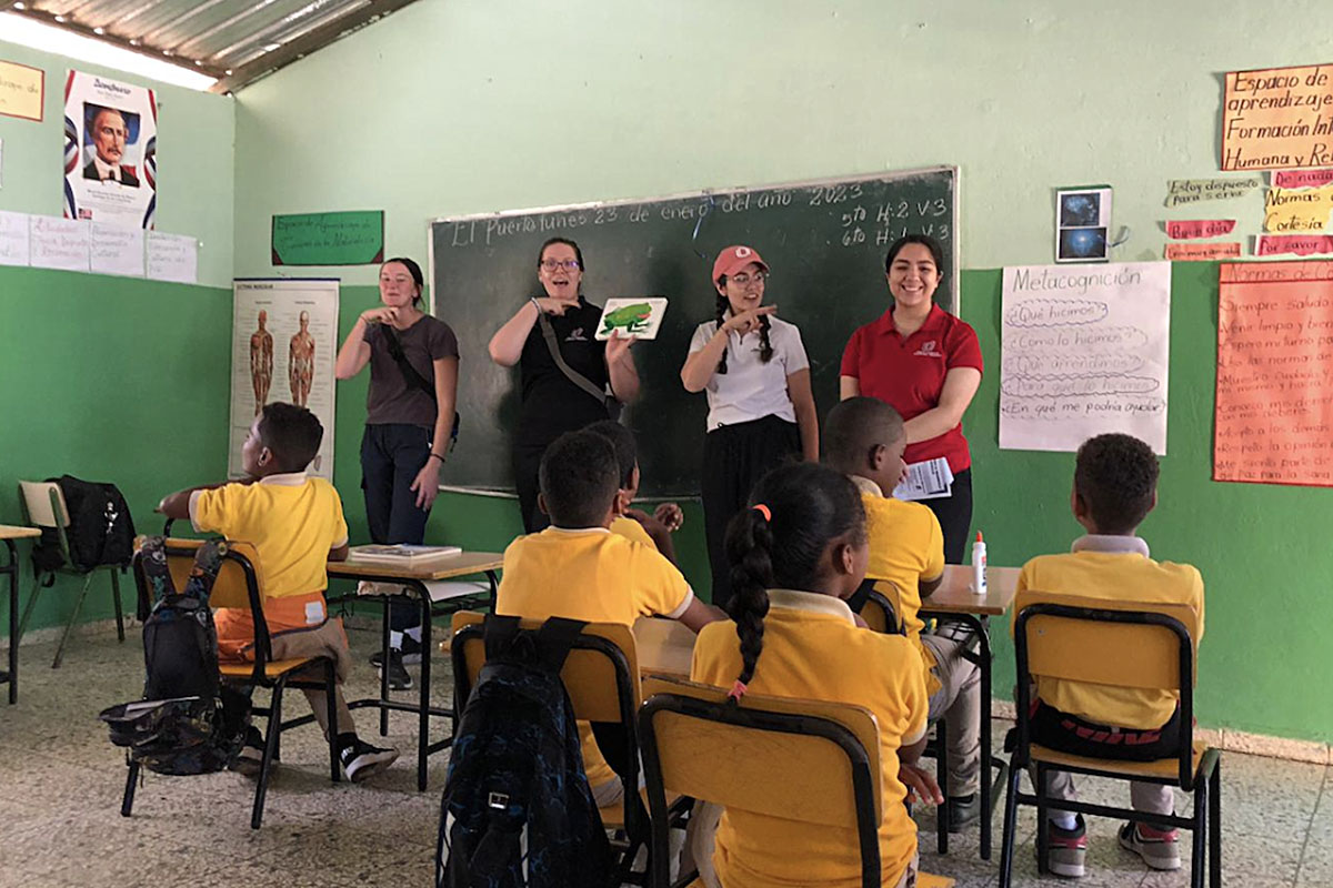 Four students present in front of a classroom to elementary-age children sitting at desks