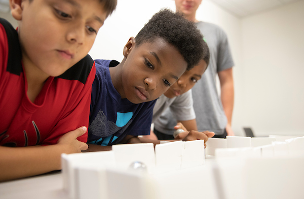 A teacher stands in the background while two students look at a project on a table