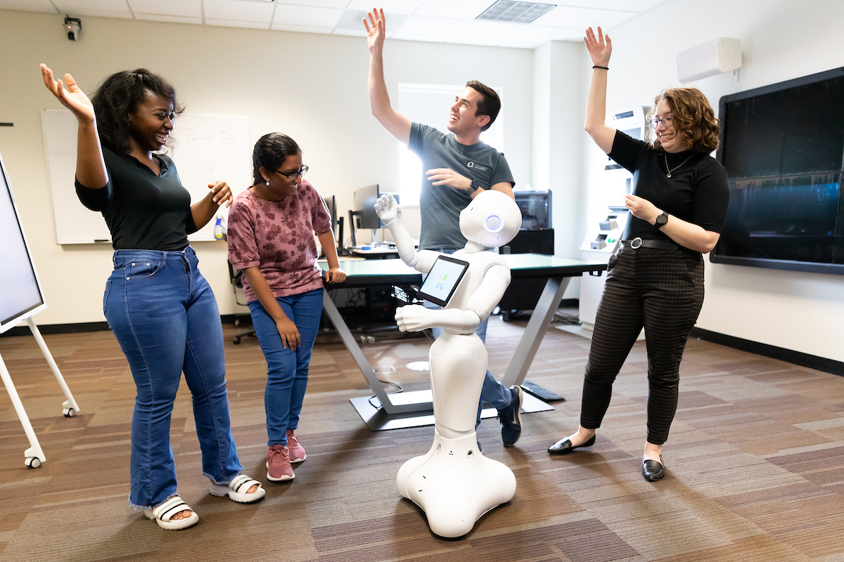 Four people and one robot each with one hand in the air while laughing.