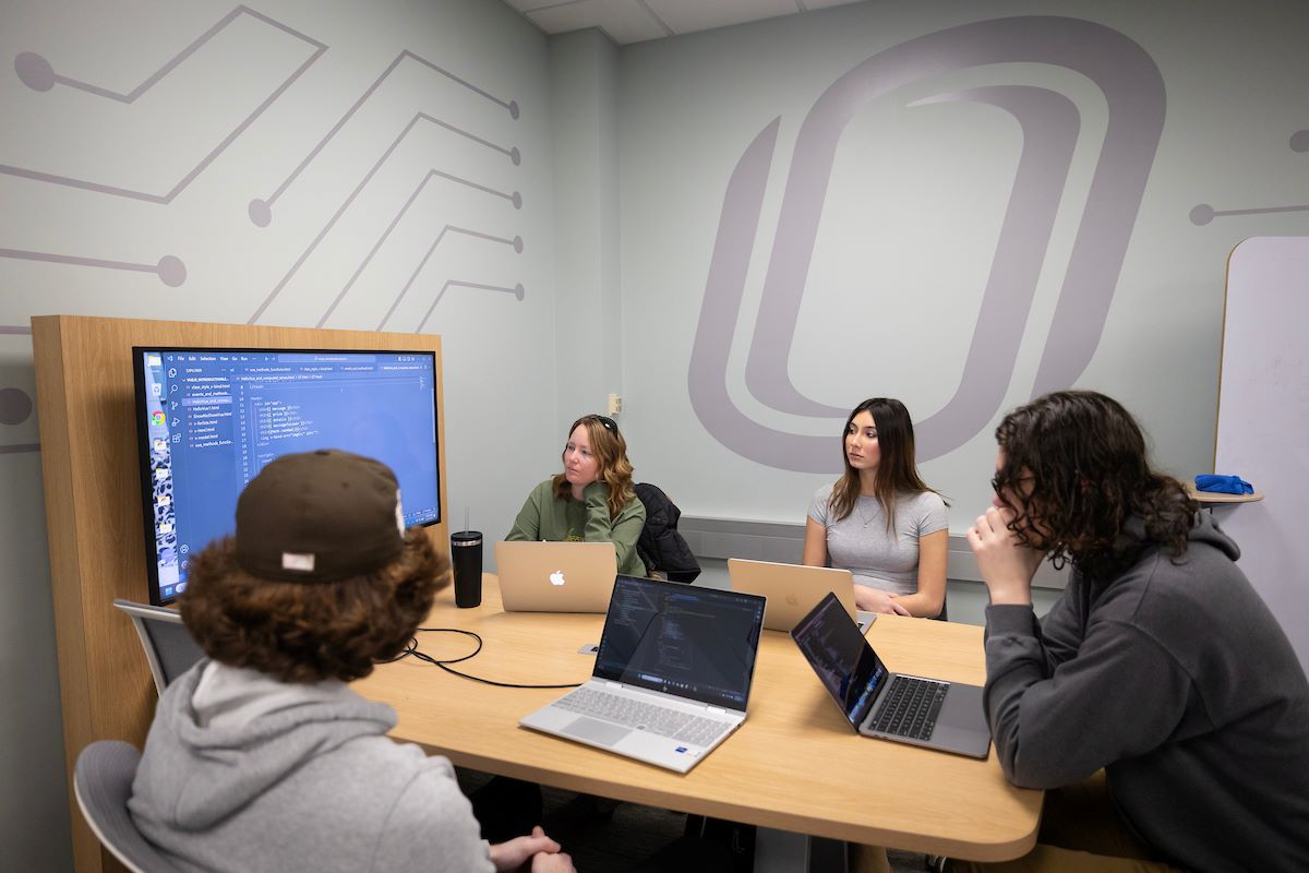 Five students sitting around a conference table while debugging code on a television.