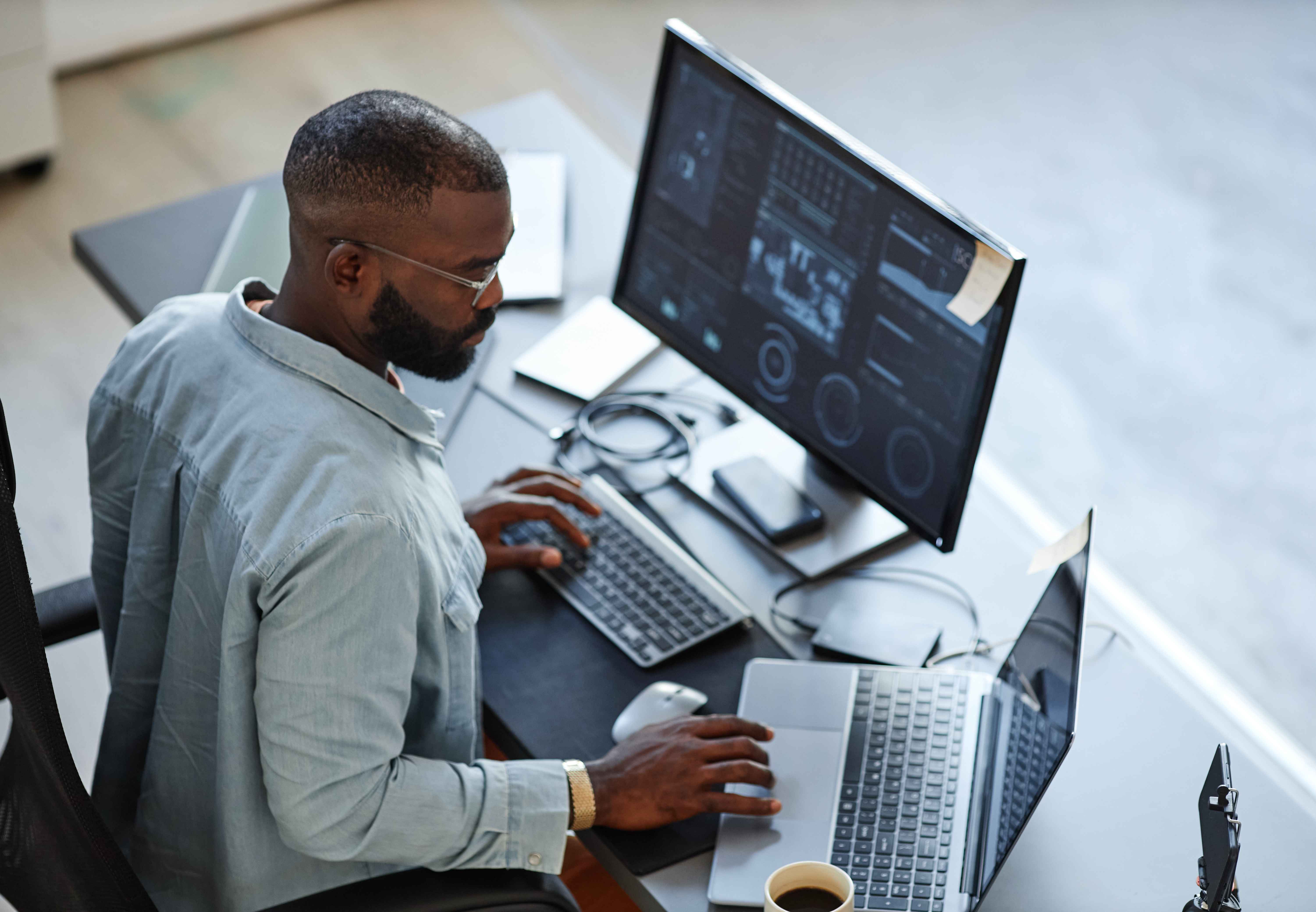 Person working at a desk with a large monitor showing data visualizations and code, alongside a laptop, notebook, phone, and coffee cup.