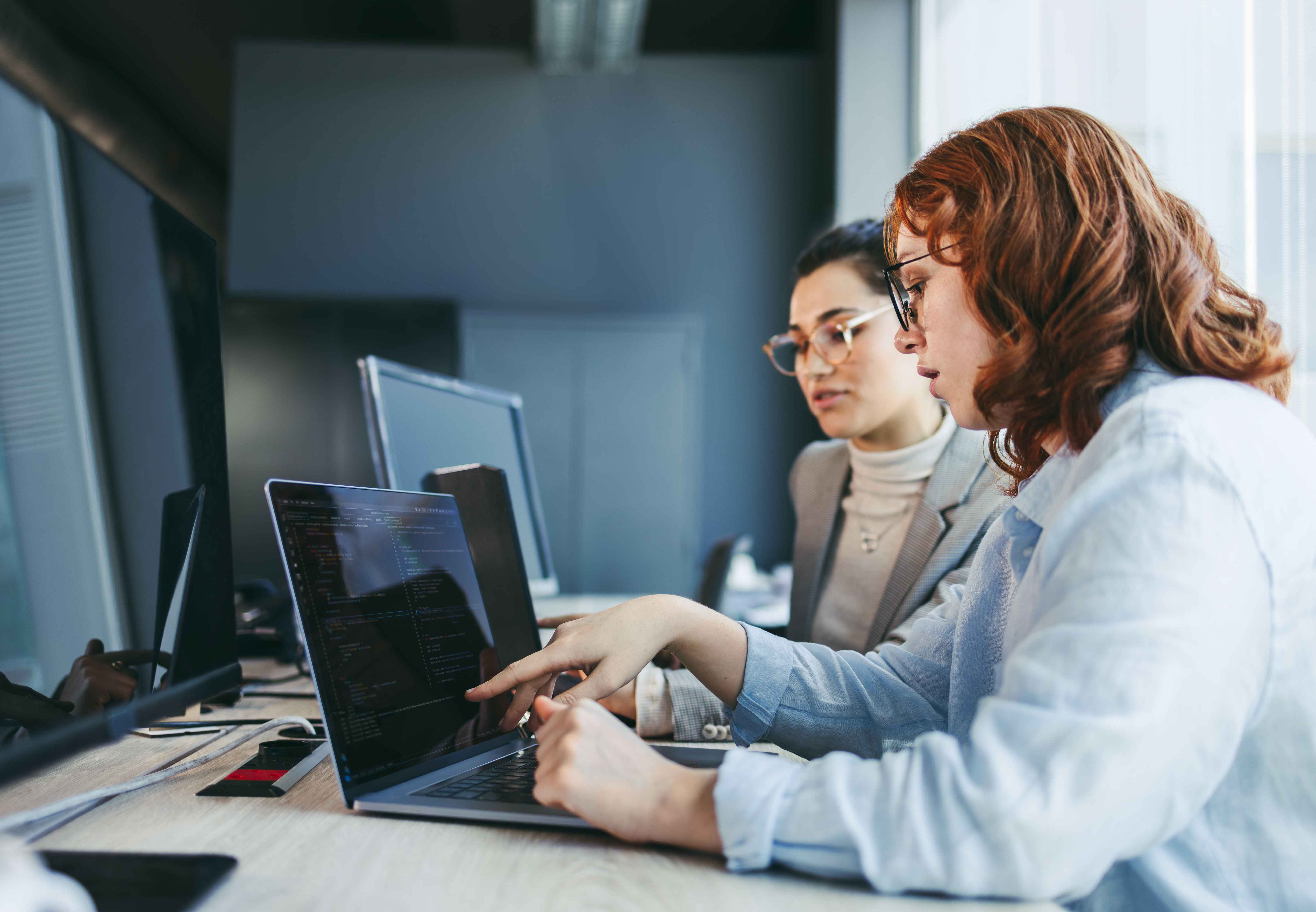 Two people in an office reviewing code on a laptop screen, with additional monitors and equipment on the desk.