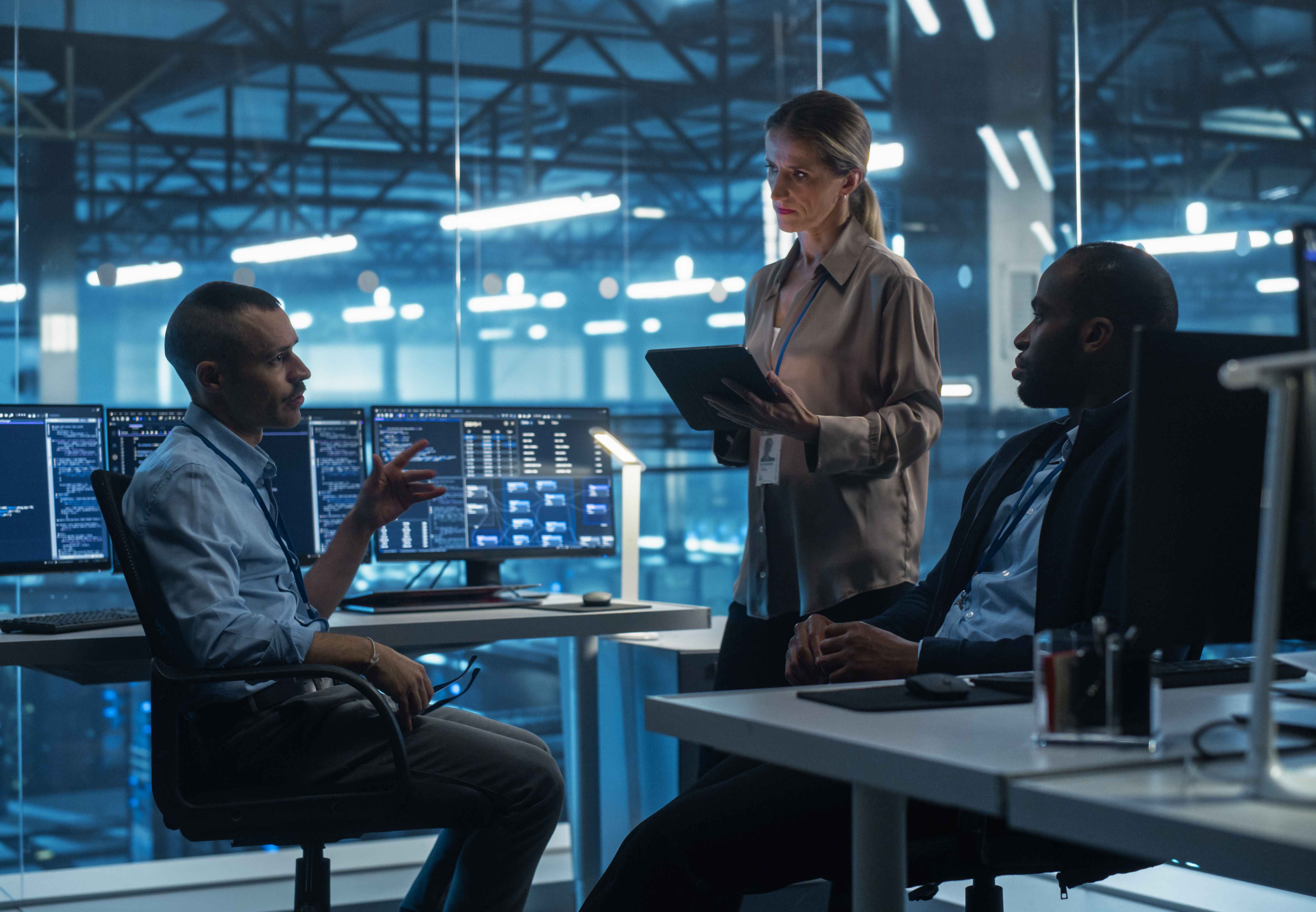 Three people in a high‑tech office with multiple monitors showing data and code, collaborating while one person holds a tablet.