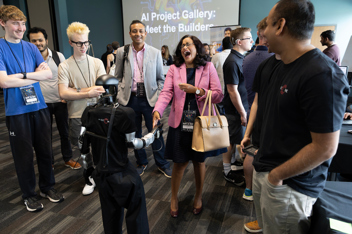Attendees laugh and interact with a robotic arm demonstration at the Mav AI Day 'AI Project Gallery: Meet the Builders' event, as a group gathers around a table-mounted robot.