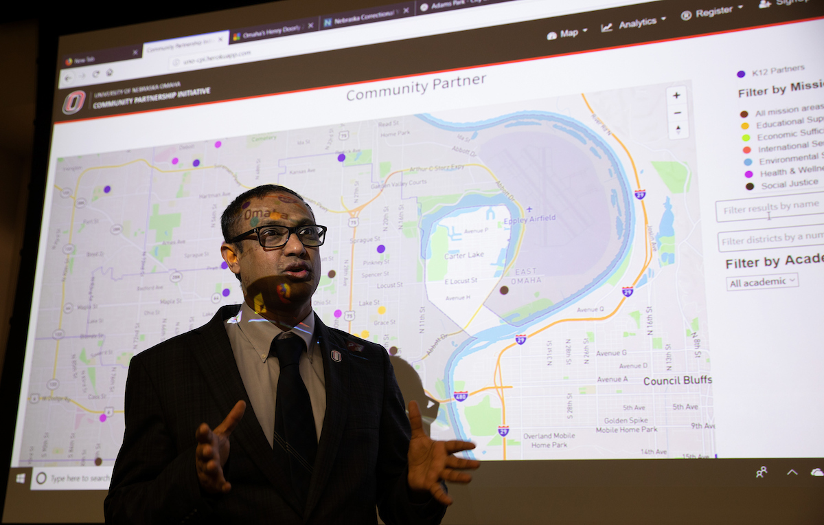 A man in a suit stands and speaks in front of a projected screen displaying a detailed map of Omaha and Council Bluffs.