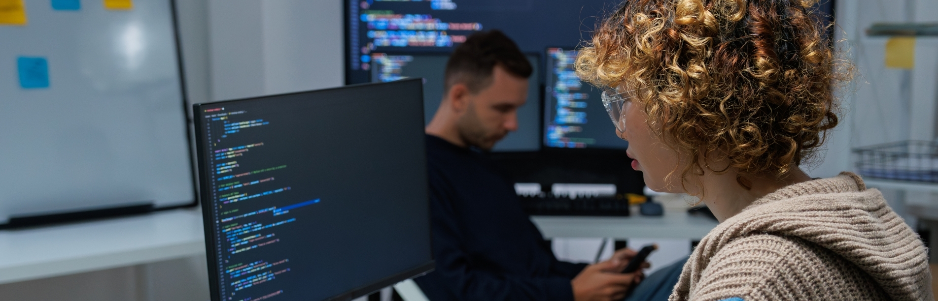  A person with curly hair and glasses works at a computer displaying colorful, syntax‑highlighted code, while another person sits farther back checking a phone beside a large monitor showing more code.