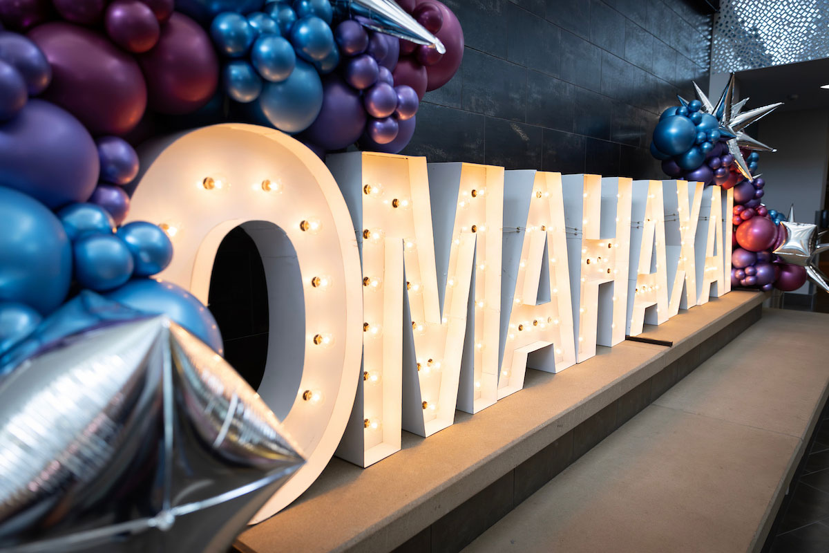 Large illuminated marquee letters spelling "OMAHAXAI" are displayed indoors, surrounded by clusters of blue, purple, and silver balloons and metallic star-shaped balloons.