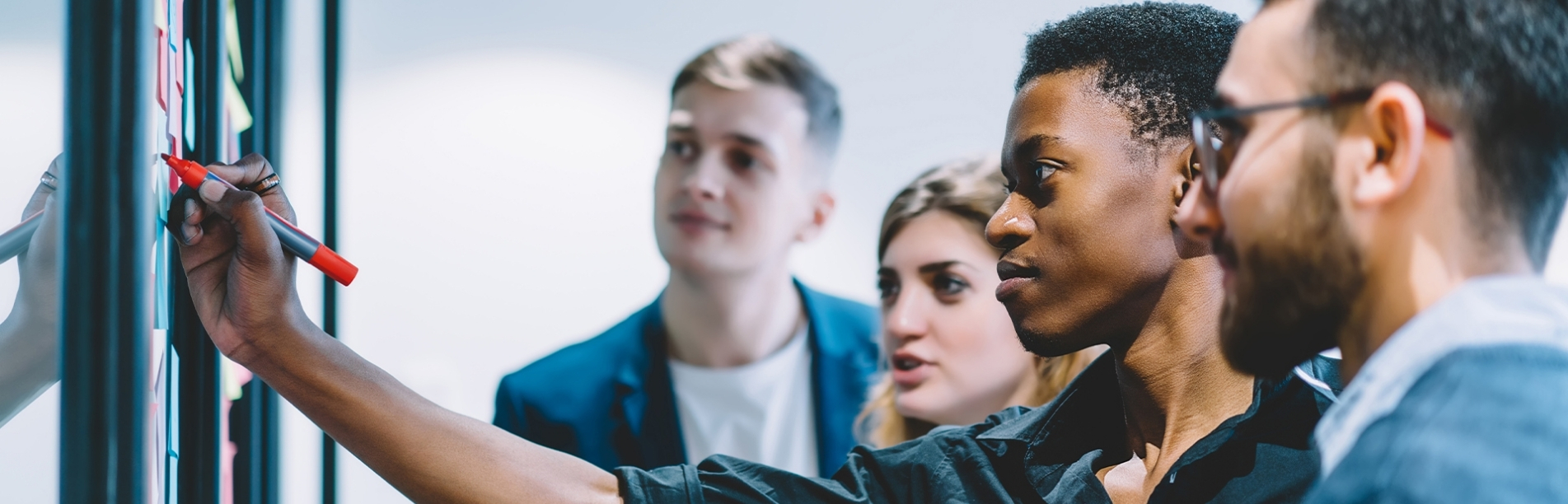 The image shows four management information systems students, a women and three men, standing in front of a white board scattered with multi-colored sticky notes. The African American man has his arm extended while holding a red dry erase marker while the others watch.