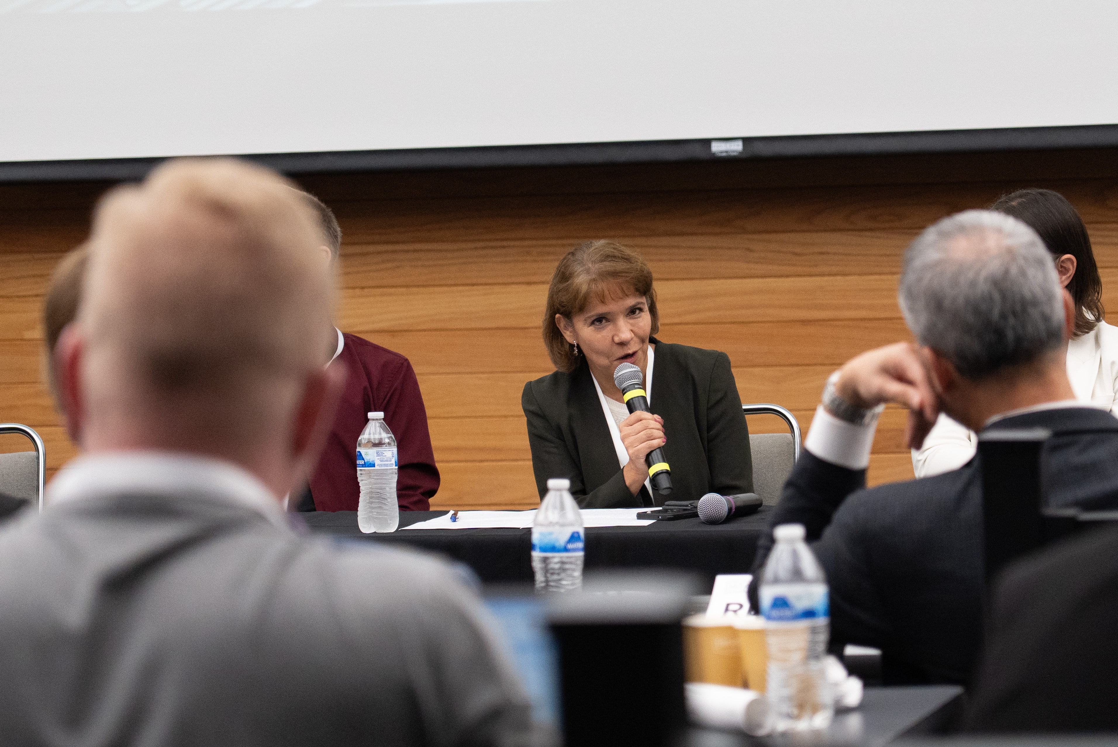 Dean Martha Garcia-Murillo speaks into microphone during panel discussion at conference table with water bottles, other panelists and audience members visible.