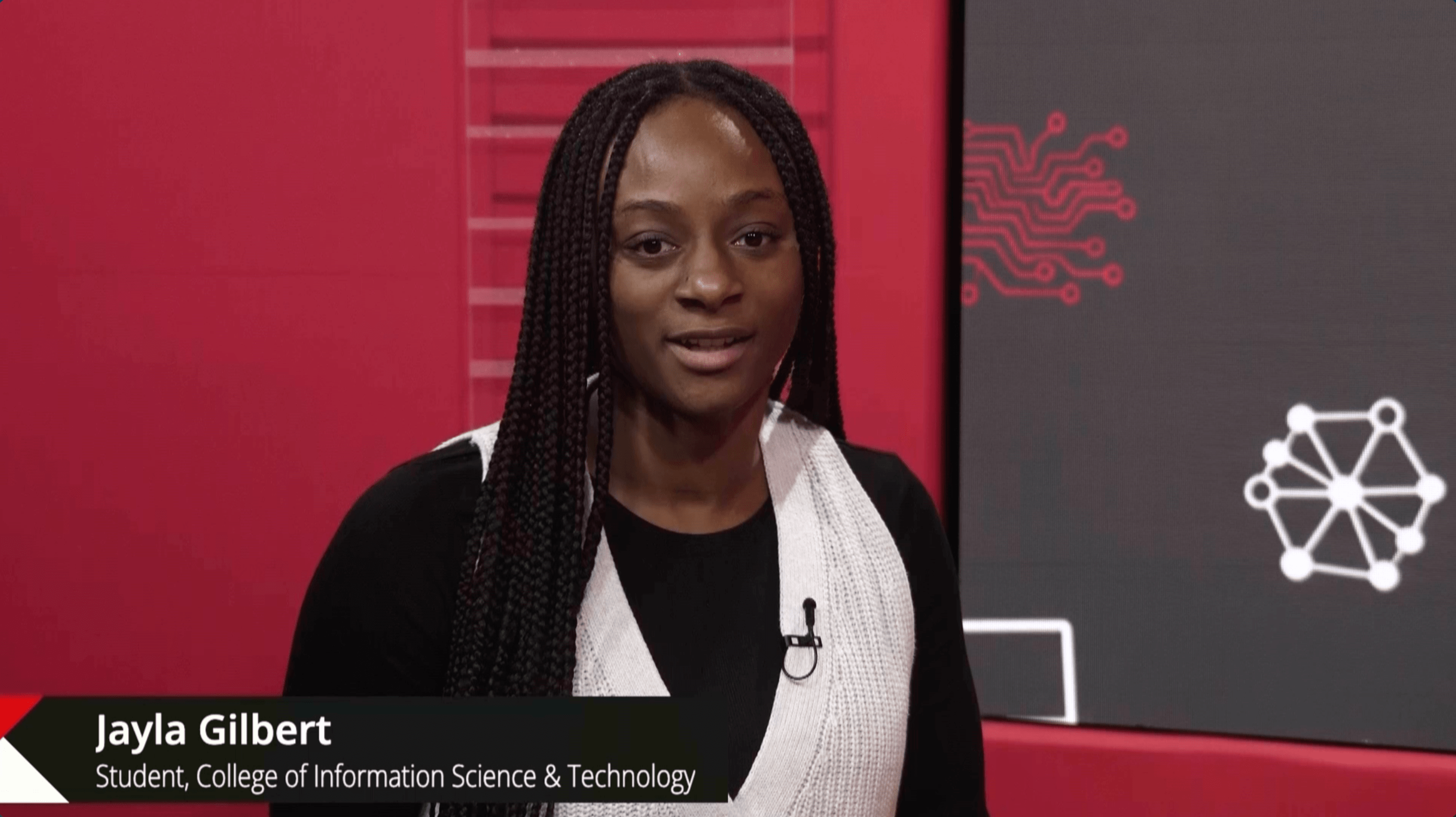 Jayla Gilbert, a student at the College of Information Science & Technology, speaks during an interview. She wears a black top with a white cardigan and has a microphone clipped to her collar. The background features red and gray panels with circuit board design elements.