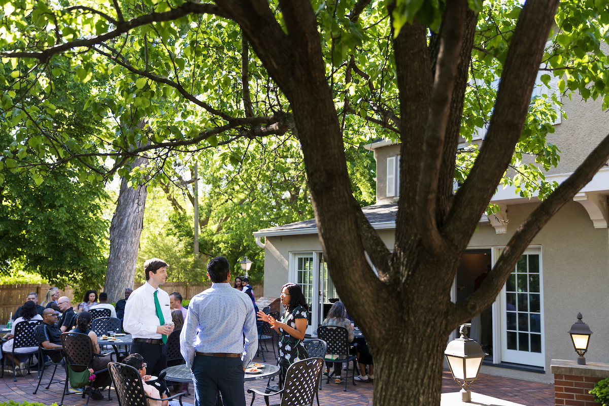 A lively outdoor gathering on a brick patio beneath large shade trees, with groups of people mingling and dining at cast iron tables near a house.