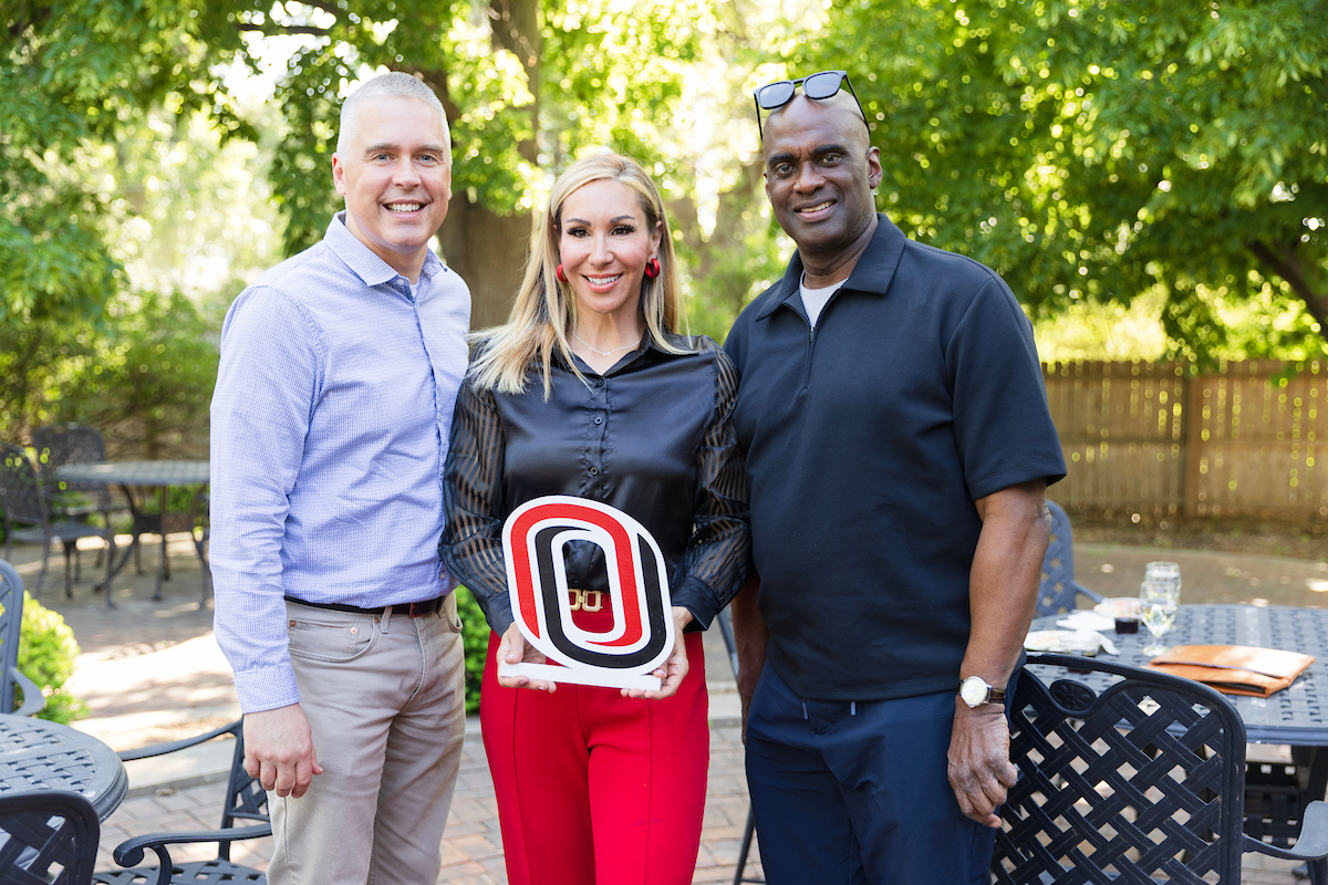 Three people posing together outdoors, smiling. The woman in the center holds a large University of Nebraska Omaha (UNO) logo prop in red, black, and white.
