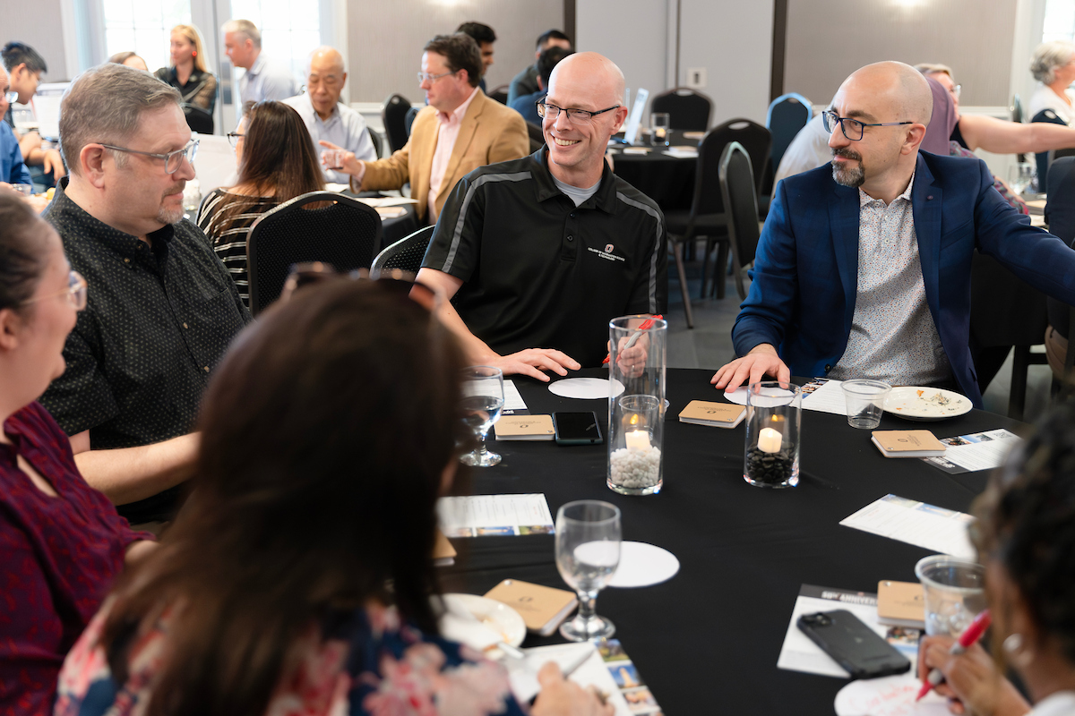 A large group of people seated at round tables with black tablecloths at an indoor event, engaged in animated conversation. Several attendees smile and interact across the table.