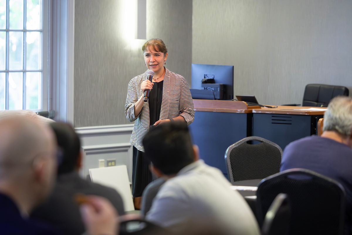 A woman in a striped blazer stands at the front of a conference room holding a microphone, presenting to a seated audience.