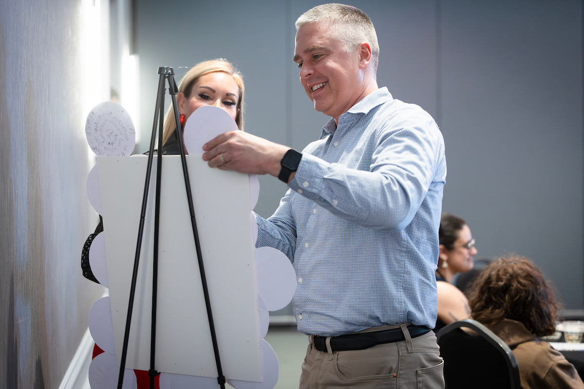 A man in a light blue shirt smiles while working at a display easel with white circular cutouts, with a woman visible behind him and other attendees in the background.