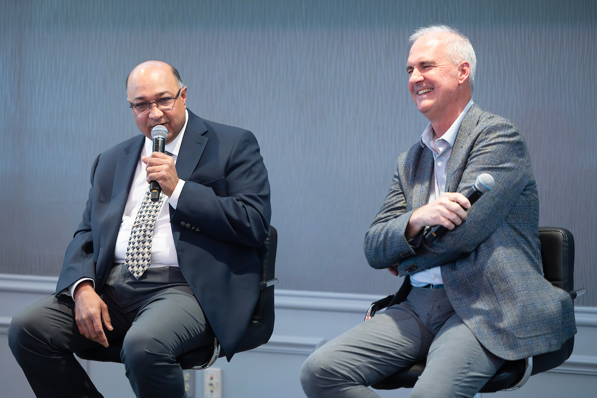 Two men seated on a panel stage, each holding a microphone. The man on the left speaks while the man on the right smiles and listens.