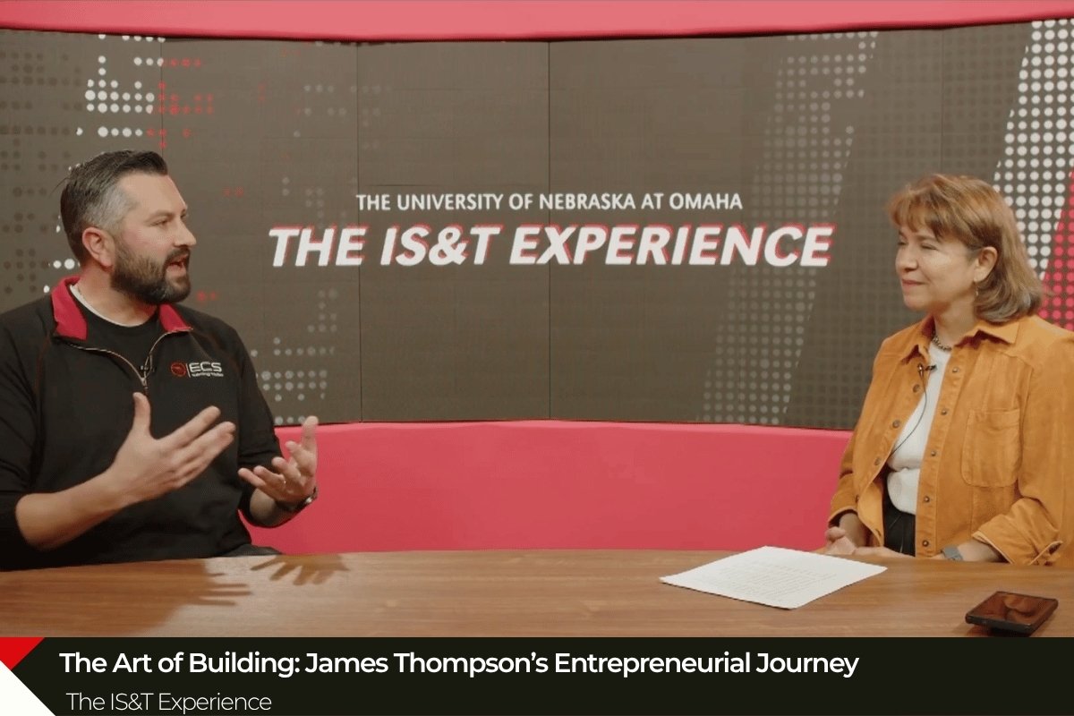Photograph of a studio interview featuring two people seated at a table with a large screen behind them displaying "The University of Nebraska at Omaha THE IS&T EXPERIENCE," with a caption at the bottom reading "The Art of Building: James Thompson’s Entrepreneurial Journey" and "The IS&T Experience."