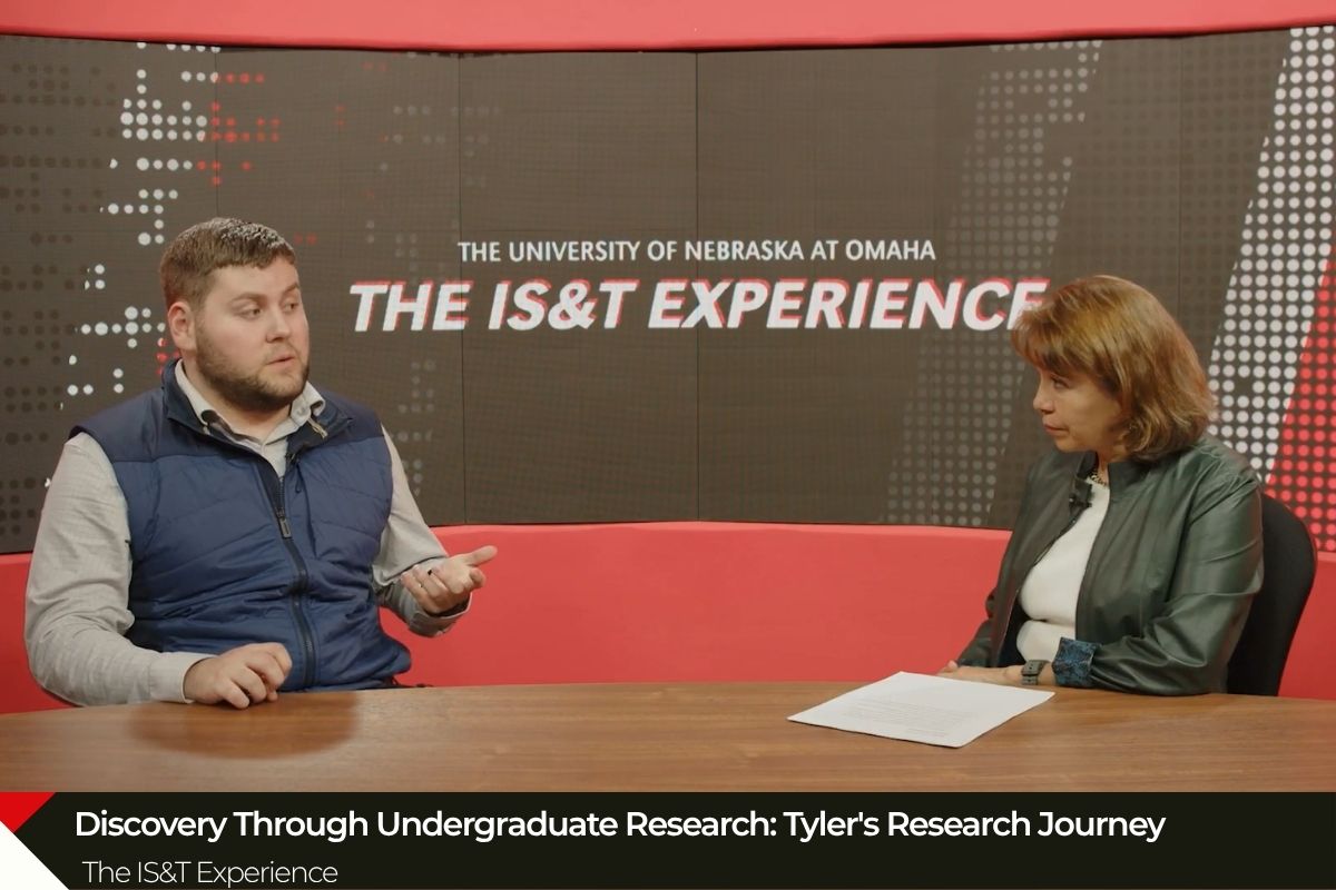Tyler McCoid (left) and College of IS&T Dean Martha Garcia-Murillo, Ph.D. (right) seated at a table in a studio setting with a large screen behind them displaying "The University of Nebraska at Omaha THE IS&T EXPERIENCE." The interview discussion focused on undergraduate research, specifically Tyler's research journey.